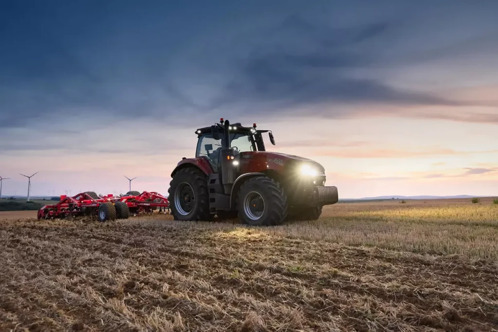 Case IH Magnum 340-traktor med arbeidslys på, kjører gjennom et jorde i solnedgang med tilkoblet redskap.