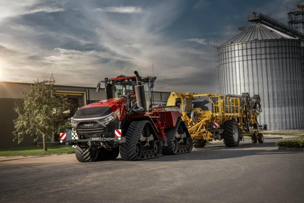 Case IH Quadtrac traktor med beltedrift foran en stor kornsilo og en gul såmaskin festet bak.