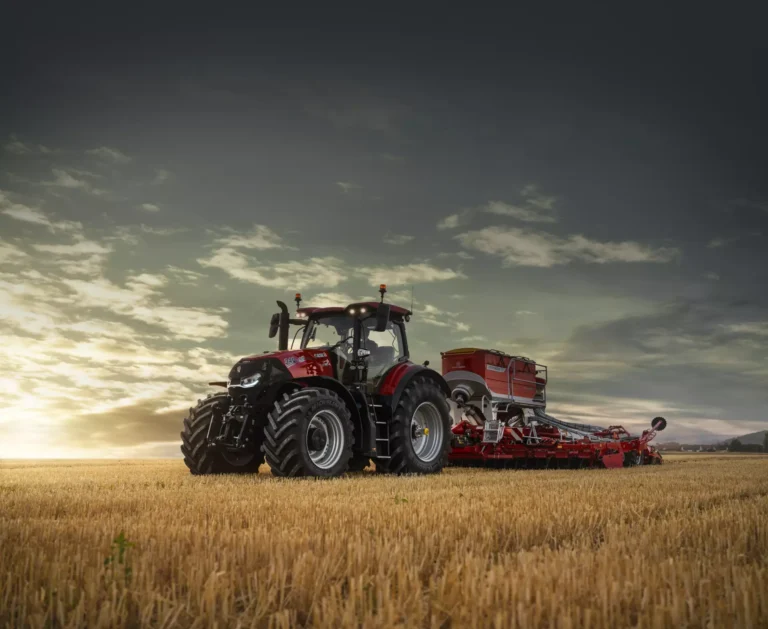 En kraftig rød traktor fra Magnum-serien står på en gyllen kornåker i solnedgangen, med en tilhenger for jordbruk festet bak. Den dramatiske himmelen og åpne landskapet fremhever traktorens robuste design og styrke.