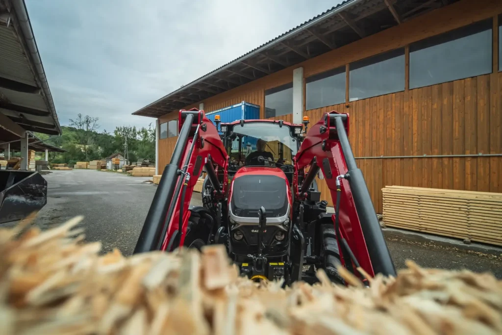 Case IH Farmall 120C-traktor med frontlaster sett bakfra, med en stor mengde treflis i forgrunnen og et sagbruk i bakgrunnen.