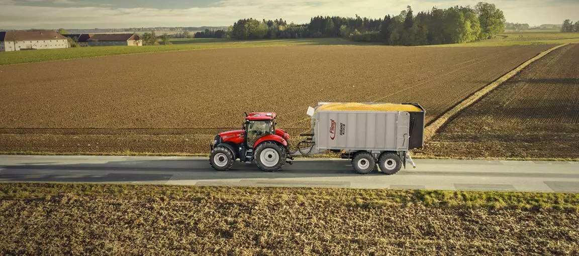 Case IH Maxxum traktor trekker en tilhenger full av avling langs en landevei med åkre på begge sider.