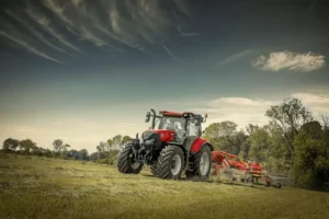 En Case IH-traktor med slåmaskin, parkert på et jorde omgitt av trær og en himmel med spredte skyer.