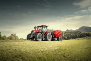 En Case IH-traktor koblet til en rundballepresse, plassert på en eng med fjell og skog i bakgrunnen.