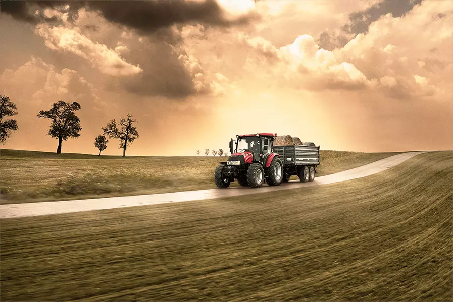 Case IH Farmall 75A traktor kjører på landevei med tilhenger lastet med høyballer.