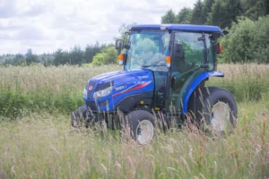 Alt-tekst: En blå Iseki TG 6507 kompakttraktor med luksushytte står i et frodig, grønt landskap omgitt av høyt gress og trær. Traktoren viser sin robusthet og passer perfekt for landbruksoppgaver under nordiske forhold.