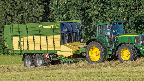 John Deere traktor med påhengskjørt i grønt og gult, fotografert under drift på åker med kornfelt i bakgrunnen.
