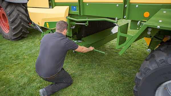 Tekniker justerer en arm eller komponent på understellet av en grønn KRONE AX rotorballe mens maskinen står på gress.