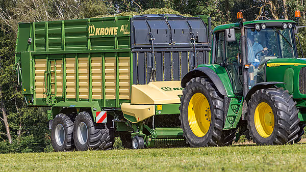 KRONE AX fôrvogn trukket av grønn traktor på grønt område, med to aksler og grønt samt gult utvendig design.