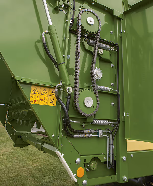 Bakside av KRONE vogn som viser hydraulisk drivverk med kjeder, sprocket-hjul, fjærer og sikkerhetsskilt for høy belastning.