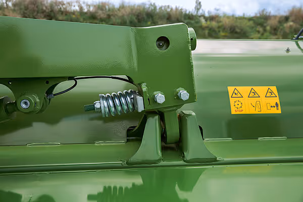 Nærbilde av fjæringssystem og sikkerhetsskilt på KRONE slåmaskin. Grønn metallkonstruksjon med hydraulisk fjæring synlig.