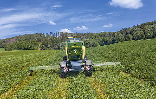 KRONE Big M 450 slåttemaskin arbeider på grønt jorde i et kupert landskap med skog og blå himmel i bakgrunnen.