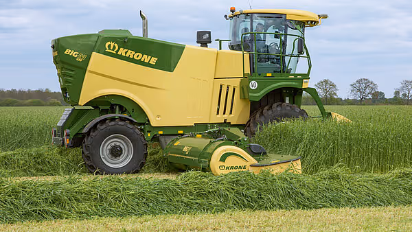 En grønn og gul Krone Big M 450 slåtterkondisjonerer arbeider i en eng og kutter gress. Maskinen har et kabinerte førerhus og store hjul som er tilpasset markarbeid.