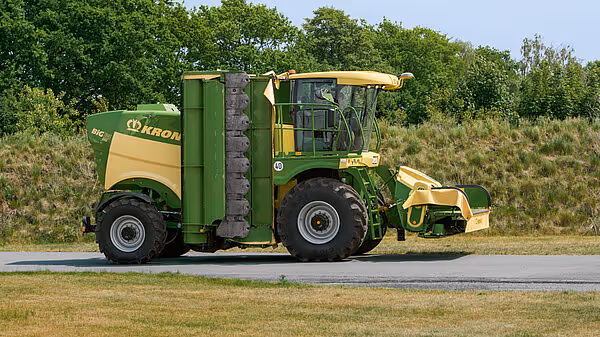 En KRONE Big M 450 selvgående slåmaskin står på en vei med grønne marker i bakgrunnen. Maskinen har grønn og gul farge og er utstyrt med slåaggregat foran.
