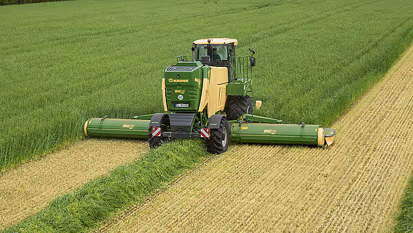KRONE Big M 450 slåmaskin i arbeid på et grasareal, sett fra lufta. Maskinen slår gras og etterlater seg tydelige striper av nyslått og uslått gras.