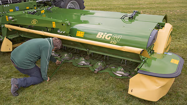 En person utfører vedlikehold på en grønn KRONE Big M 450 slåmaskin som ligger på bakken. Maskinen har gule beskyttelsesdeksler og personen arbeider med undersiden av utstyret.