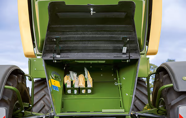 Åpen oppbevaringsboks på en KRONE Big M 450 slåmaskin som viser verktøy og reservedeler lagret inne i det gule og grønne kabinettet. Boksen er montert på maskinen for enkel tilgang under drift.