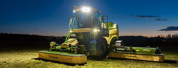 Krone Big M 450 slåmaskin med arbeidsbelysning i skumringstid på eng. Maskinen har gul og grønn fargekombinasjon med utslåtte slåtteredskap.