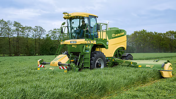 KRONE Big M 450 slåmaskin i drift på en grønn eng med skog i bakgrunnen. Maskinen slår gras og har karakteristisk grønn og gul KRONE-fargekombinasjon.