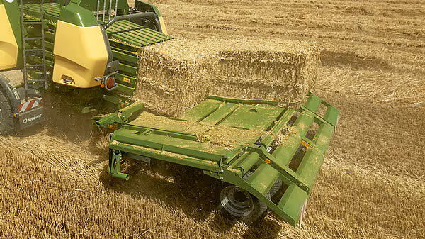 Nærbilde av en Krone BigPack storballpresse som slipper en ferdig firkantballe på en grønn samletilhenger i åkeren.