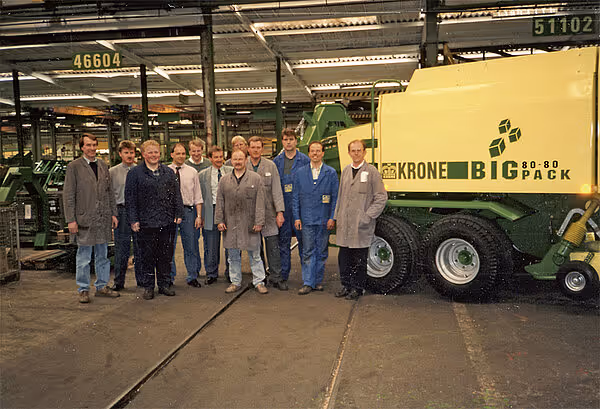 Gruppe menn i arbeidsklær står foran en grønn og gul KRONE BigPack 80-80 storballpresse i en industrihall.