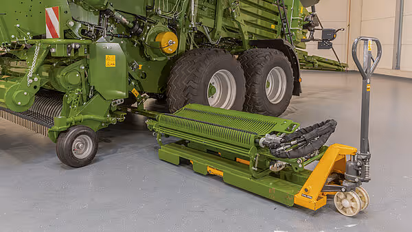 KRONE Big Pack pressemaskin med oppsamlingsvalse montert på gul hydraulisk jekk. Maskinen står i verkstedhall ved siden av hovedenheten.