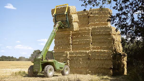 En grønn teleskoplaster løfter storballer ved siden av en høy stabel med gule storballer i et landskap med åkrer og trær.