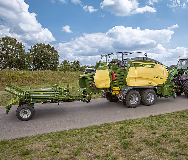 En Krone BigPack storballpresse med tilhenger transporteres på en asfaltert vei gjennom et landlig område.