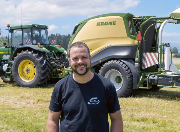 En mann i svart t-skjorte står smilende foran en grønn og gul KRONE CombiPack HDP rundballpresse og en grønn traktor på en grasmark.