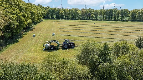Flyfoto av traktor med KRONE CombiPack HDP rundballpresse i arbeid på slått eng, med ferdige rundballer spredt utover feltet.