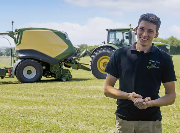 En ung mann i mørk poloskjorte står på en grasmark med en grønn og gul KRONE CombiPack HDP rundballpresse og traktor i bakgrunnen.