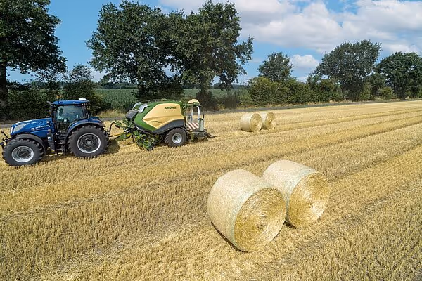 En blå traktor med gul KRONE ballepresse arbeider på en stubbåker med flere ferdigproduserte rundballer i forgrunnen.