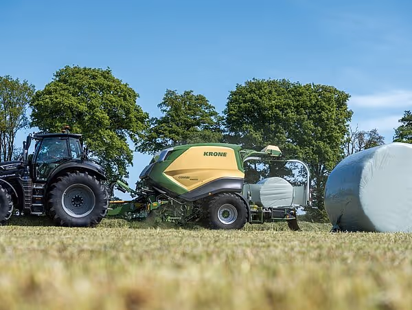 Traktor med KRONE CombiPack HDP rundballpresse på åker omgitt av grønne trær under blå himmel.