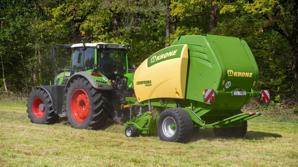 KRONE Comprima rundballpresse tilkoblet grønn traktor med røde felger under høyslått på eng ved skogkant.