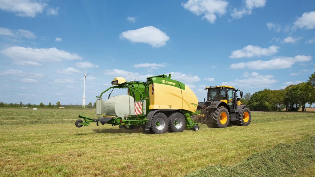 KRONE Comprima Plus rundballpresse i arbeid på eng, trukket av gul traktor med ferdigpressede rundaller spredt utover jordet.