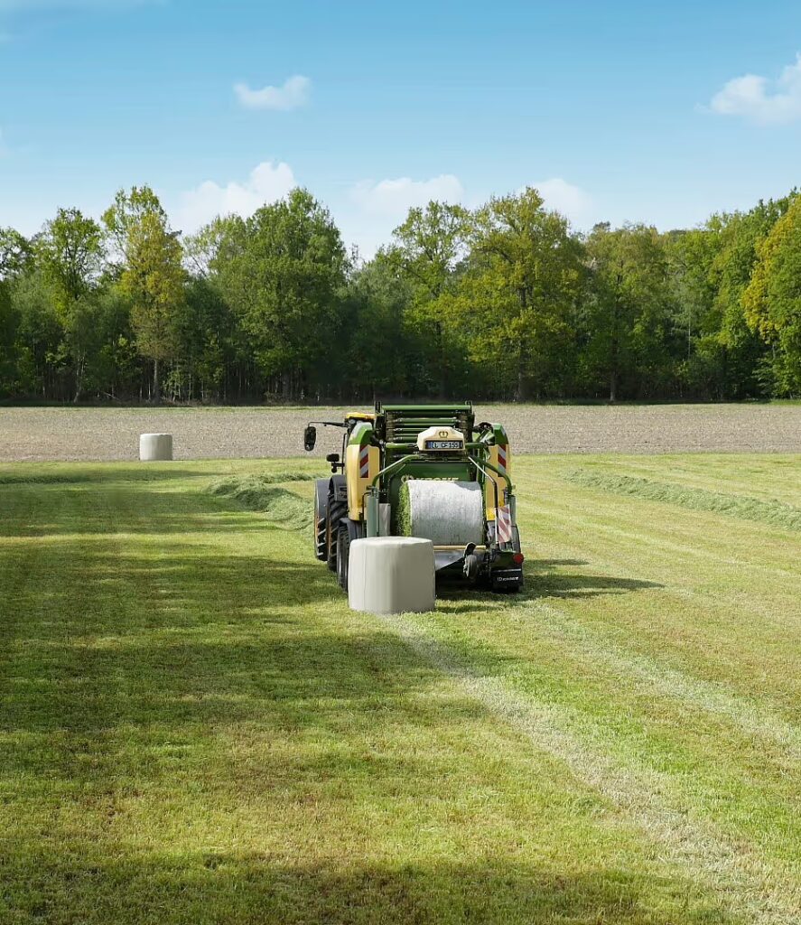 Rundballpresse som produserer hvite plastinnpakkede rundaller på grønn eng med skog i bakgrunnen.