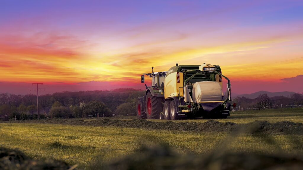 KRONE Comprima Plus rundballpresse arbeider på åkeren i solnedgangen. Maskinen er koblet til traktor og pakker høy eller halm til store rundballer.