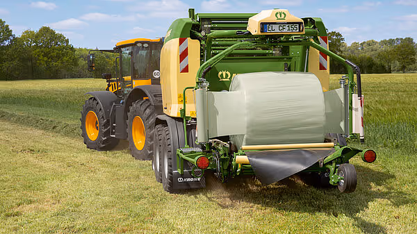 En grønn KRONE Comprima Plus rundballpresse trekkes av en gul traktor over en eng med ferdig kuttet gress.