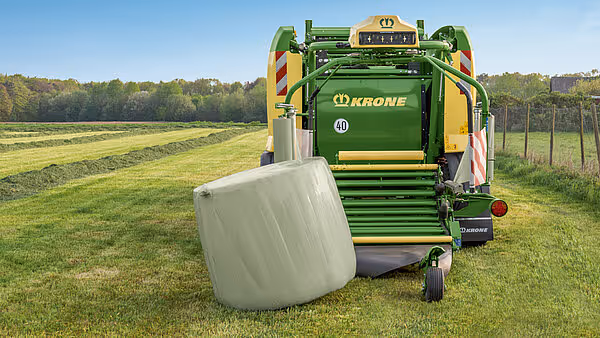 KRONE Comprima rundballpresse produserer en innpakket rundball på en grønn eng. Maskinen har karakteristisk grønn og gul farge.