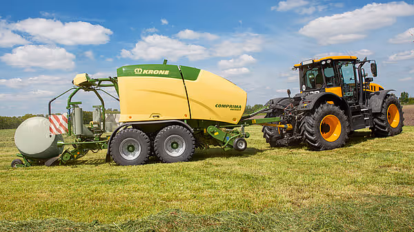KRONE Comprima rundballpresse trukket av en traktor arbeider på en slått eng under blå himmel. Maskinen pakker høy i hvit plast.
