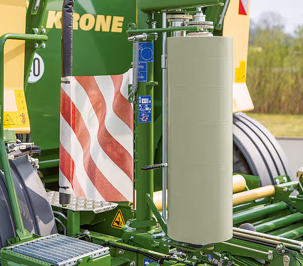 Nærbilde av folieviklingsmekanismen på KRONE rundballpresse med rød og hvit sikkerhetsskilt. Viser den tekniske løsningen for automatisk folieomvikling.