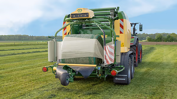 KRONE Comprima Plus rundballpresse sett bakfra på grønt jorde. Maskinen produserer hvite plastinnpakkede rundballer og har gul og grønn fargekombinasjon.