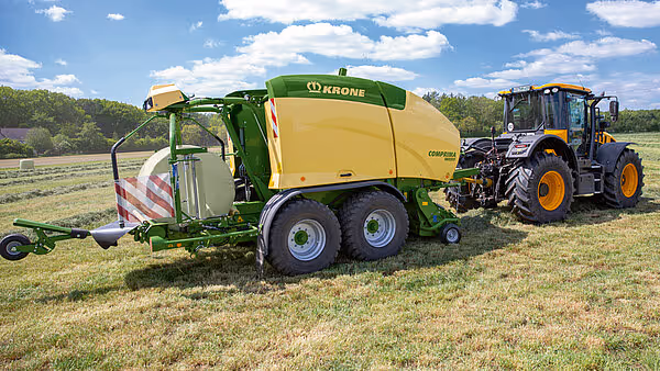KRONE Comprima rundballpresse arbeider på slåttemark, trukket av gul traktor under blå himmel.