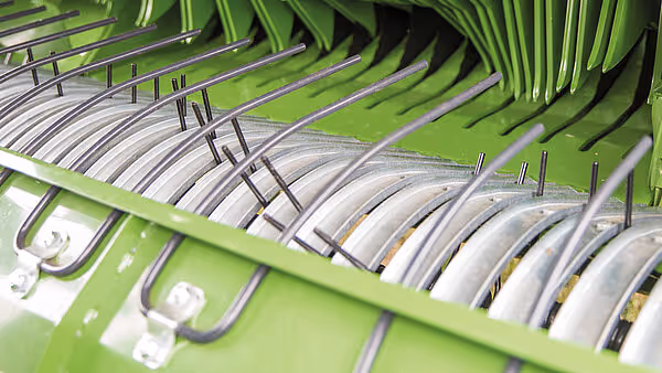 Detaljbilde av oppsamlingsenheten på KRONE Comprima med metalliske tiner som løfter høyet inn i pressen. Tinene er arrangert i rader på den grønne rammen.