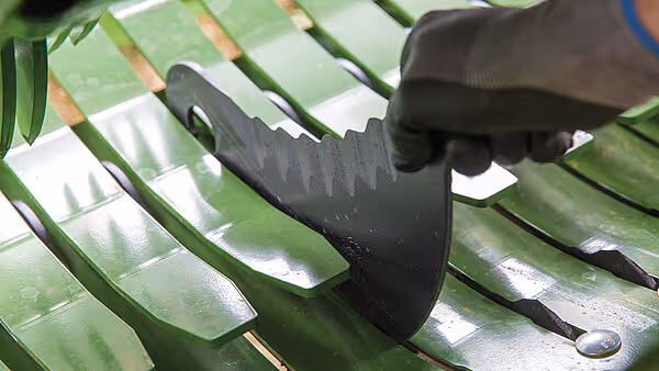 Hånd med svart hanske holder et sort plastknivblad mot grønne metallplater på KRONE Comprima rundballpresse. Kniven brukes til kutting av materiale.