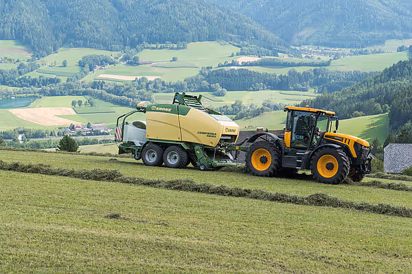 KRONE Comprima rundballpresse trukket av gul traktor på slåttemark i dallandskap.