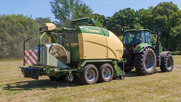 KRONE Comprima rundballpresse med wrapper-funksjon trukket av grønn traktor, maskinen har plastfolieruller for innpakking av rundaller.