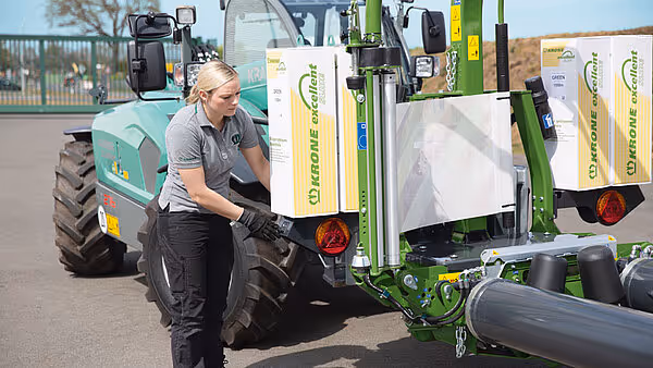 Kvinnelig operatør i grå arbeidsklær står ved KRONE EasyWrap 150 ballpresse med hvite plastfolieruller montert på siden.