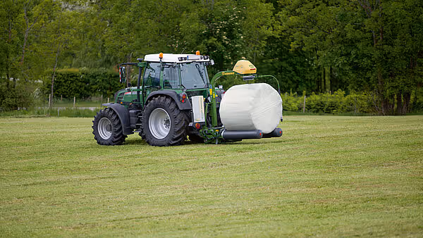 Grønn traktor med påmontert KRONE EasyWrap 150 rundballepresse som produserer hvite plastinnpakkede rundballer på slåttemark.
