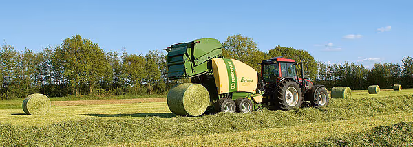 Krone Fortima rundballpresse i arbeid på åker, produserer store rundaller mens den trekkes av grønn traktor.