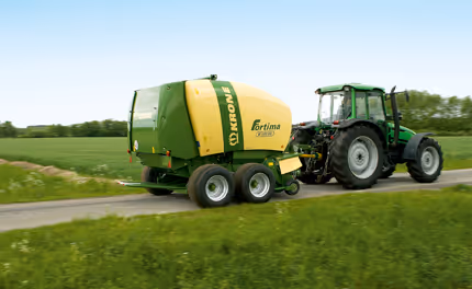 Krone Fortima rundballpresse transporteres på vei av grønn traktor, viser maskinen i transportposisjon.